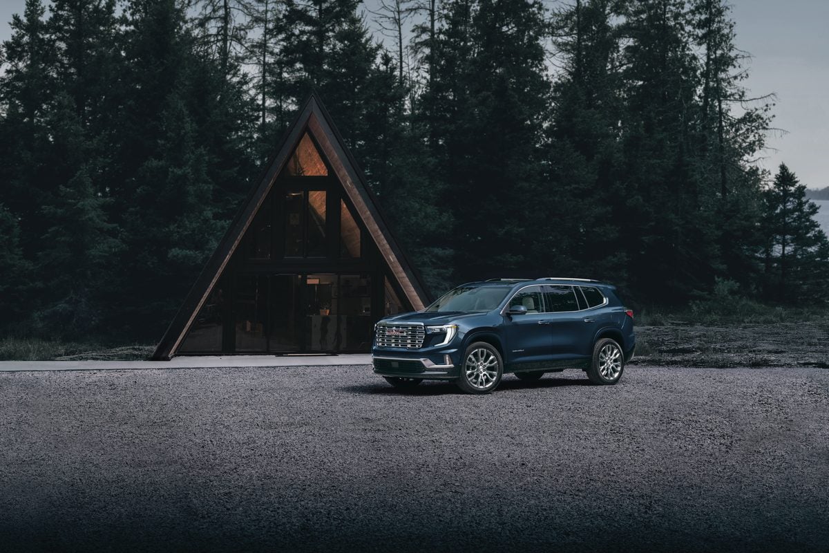 Blue 2027 GMC Acadia Denali parked in front of an A-frame cabin among tall pines