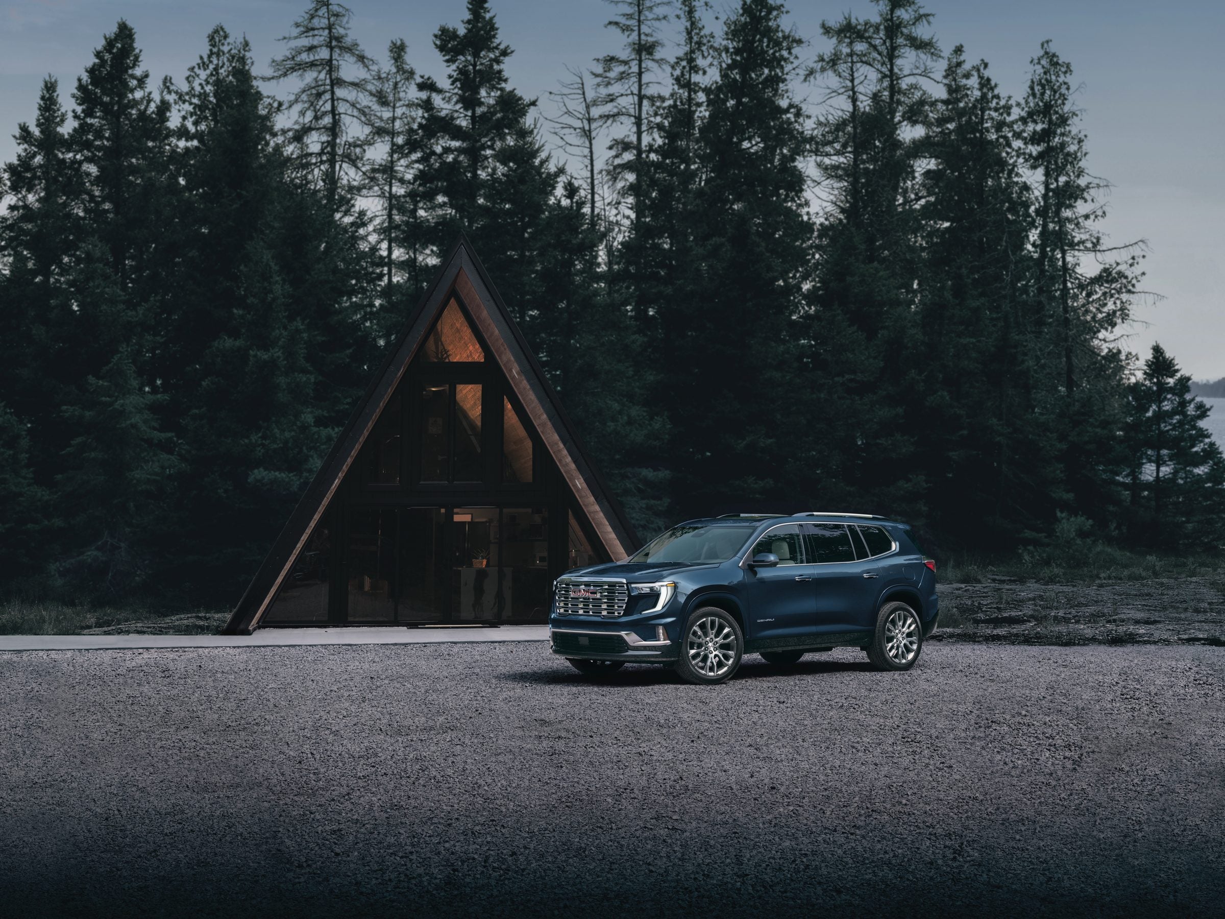 Blue 2027 GMC Acadia Denali parked in front of an A-frame cabin among tall pine trees
