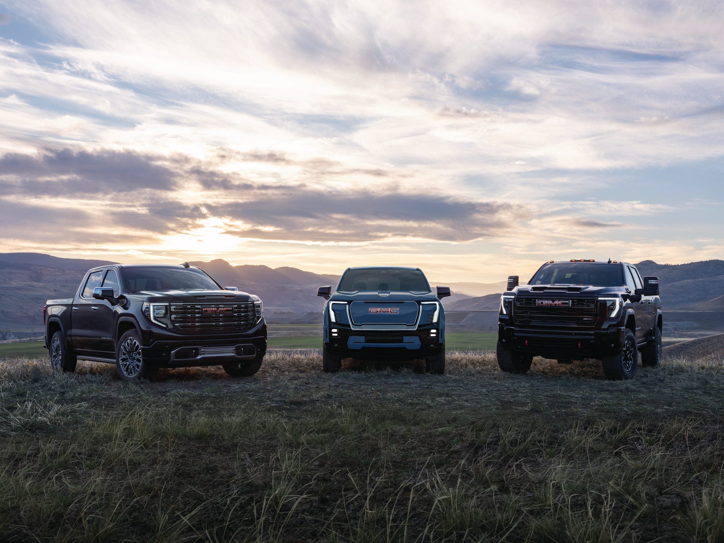 Three GMC trucks lined up on a grass ridge at sunset, a 2027 model year lineup shot