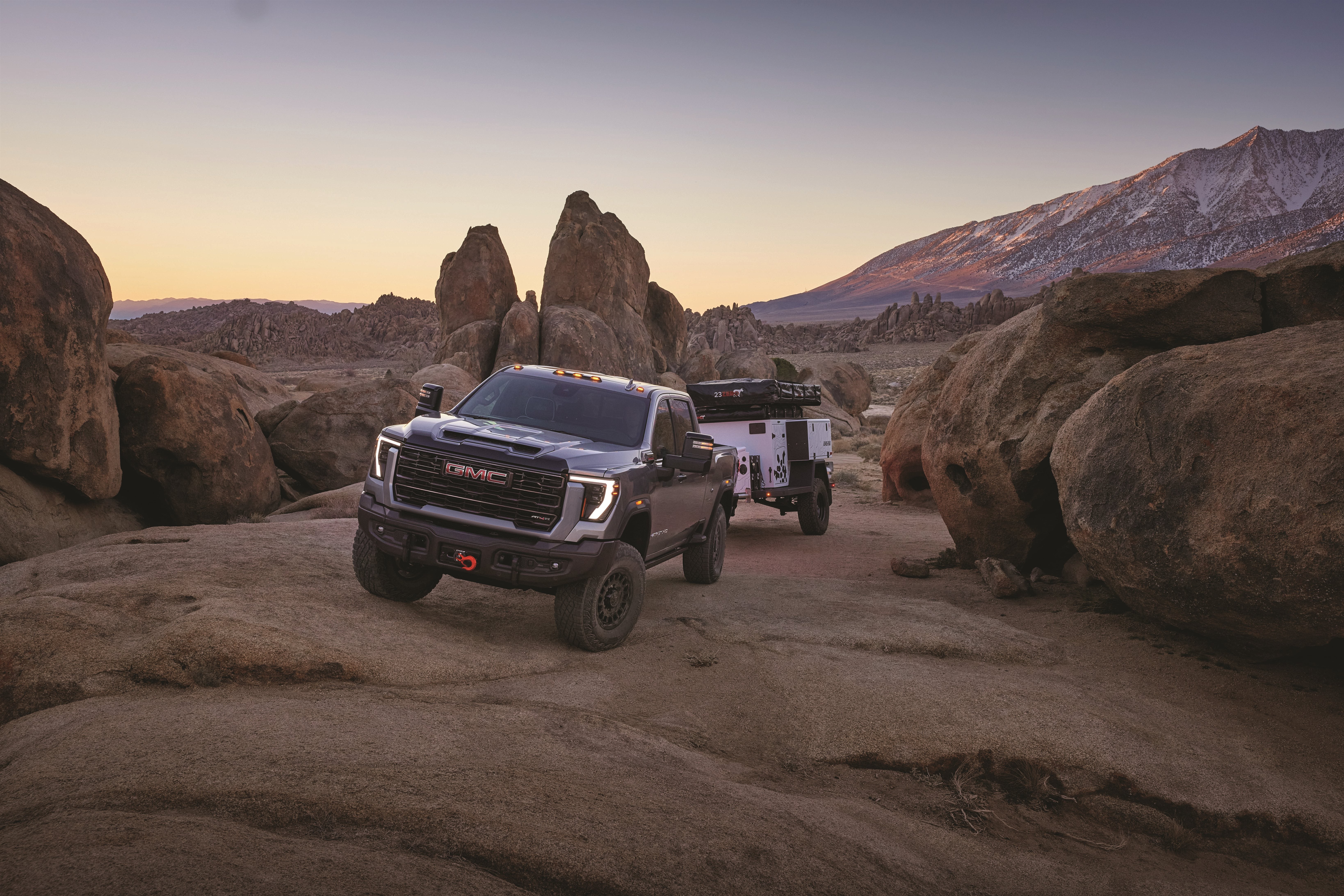 2027 GMC Sierra HD AT4X towing an off-road trailer through desert rock terrain at dusk